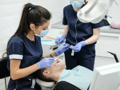 Dwie dentystki w maseczkach i rękawiczkach wykonują zabieg stomatologiczny u pacjenta.